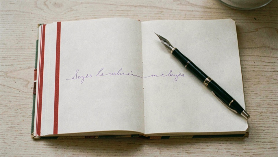 Un cahier à réglure Seyès ouvert à plat sur une table en bois clair, la marge rouge verticale bien visible à gauche, une ligne d’écriture cursive à l’encre violette tracée à moitié, un stylo plume posé en travers de la page.