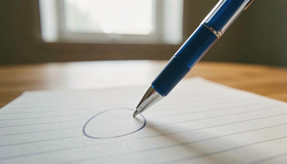 Une bande de modèle d’écriture cursive collée en haut d’un cahier, les lettres liées tracées à l’encre bleue, sous elle la même phrase recopiée par un enfant, plus grande et plus tremblée, lumière de classe.